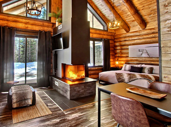 Intérieur d'un chalet en bois rond avec un foyer central et des grandes fenêtres.