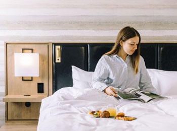 Une femme en pyjama est assise sur un lit. Elle lit un magazine et il y a un plateau déjeuner sur la douillette.