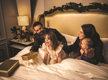 Deux parents avec une jeune fille et un bébé dans un lit d'hôtel aux allures luxueuses.