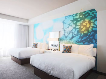 Chambre d’hôtel illuminée de lumière naturelle, avec deux grands lits, literie blanche et mur d’accent bleu.