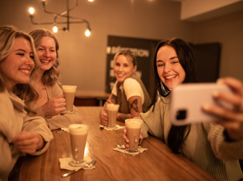 4 jeunes femmes autour d'une table qui boivent des cafés et prennent une photo de groupe.