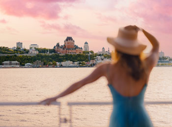 Femme en robe bleue avec un grand chapeau observant le Château Frontenac depuis le fleuve.