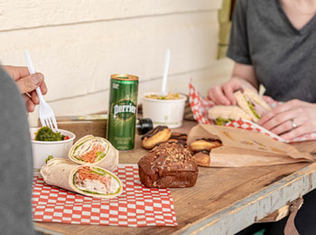 Deux personnes assises autour d’une table sur laquelle se trouve des sandwichs, des pâtisseries et d’autres items de lunch.
