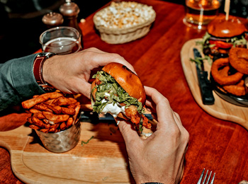 Deux mains tenant un hamburger au-dessus d'une planche avec des frites sur une table de restaurant.