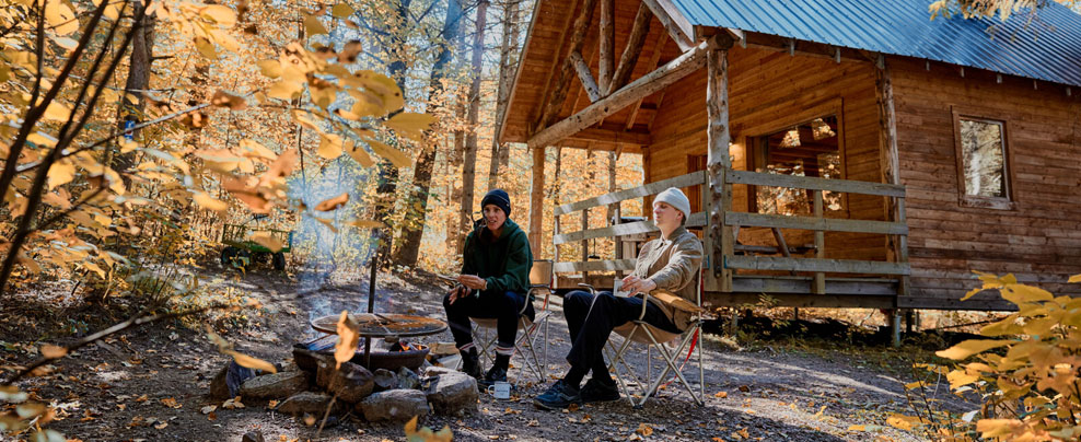 Deux personnes assises autour d’un feu de camp devant un chalet en bois dans une forêt à l’automne.