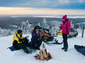 3 motoneigistes en pause autour du feu en haut d'une montagne dans les Laurentides