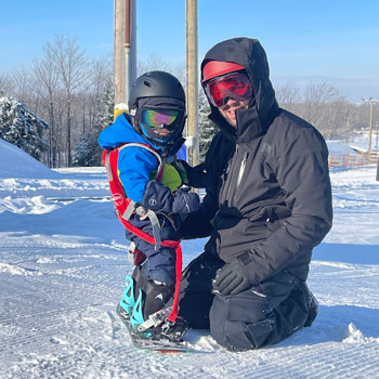 Un père avec son garçon, le bambin a une planche à neige aux pieds et le père est agenouillé devant lui.