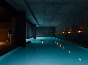 Piscine intérieure dans la pénombre, éclairée par une douce lumière; une femme se tient au bord de la piscine.