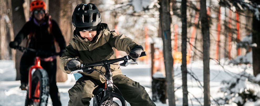 Jeune garçon en fatbike dans une forêt enneigée en avant-plan, avec une autre personne plus loin derrière.