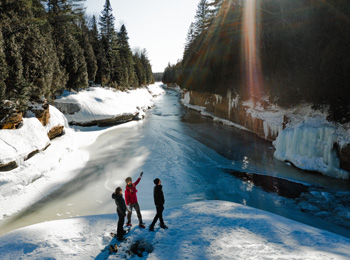 3 personnes en raquettes qui traversent une rivière dans la forêt en hiver.