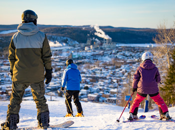 Père, fils et fille en habits de ski sur la montagne avec la vue du centre-ville devant eux.